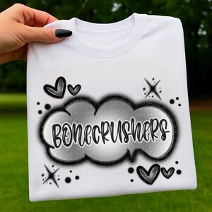 Puede incluir: Sudadera blanca de cuello redondo con la palabra "BONECRUSHERS" en un diseño de nube en blanco y negro. El diseño incluye corazones, estrellas y puntos negros. La sudadera es sostenida por una mano con una uña pintada de negro.