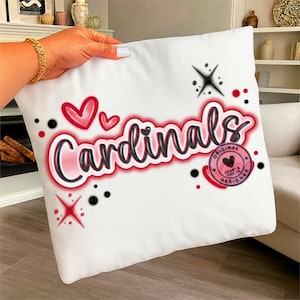 Puede incluir: Cojín cuadrado blanco con la palabra "Cardinals" en una fuente roja y negra. El cojín tiene dos corazones rojos encima de la palabra y un logotipo circular con un corazón y las palabras "Heart & Soul Designer".