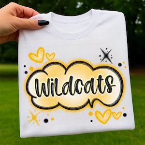 Puede incluir: Camiseta blanca con la palabra "Wildcats" en un diseño en forma de nube amarilla y negra. La camiseta presenta corazones amarillos, estrellas y puntos negros. La camiseta está sujeta por una mano.