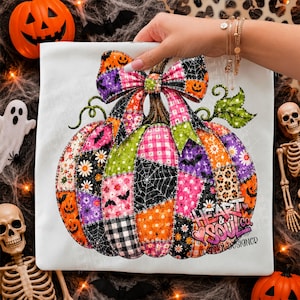 May include: A white t-shirt featuring a large, colourful patchwork pumpkin graphic. The pumpkin is composed of various patterns including orange jack-o'-lanterns, pink gingham, black spiderwebs, purple bats, and daisies, topped with a large bow.