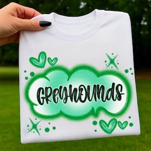 May include: White crew neck sweatshirt with the word "GREYHOUNDS" in black script inside a green cloud design. Green hearts and starbursts decorate the shirt. The sweatshirt is held up by a hand.