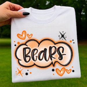 Puede incluir: Sudadera blanca con la palabra "Bears" en un diseño de burbuja negro y naranja. El diseño incluye corazones, estrellas y puntos naranjas. La sudadera es sostenida por una mano.
