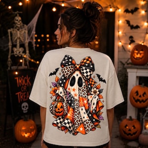 Op de afbeelding: Een crèmekleurig t-shirt met een grote afbeelding op de achterkant van een geest met een strik. De geest is versierd met oranje, zwarte en witte patronen, waaronder luipaardprint, sterren en ruitjes. De geest houdt een pompoen vast.