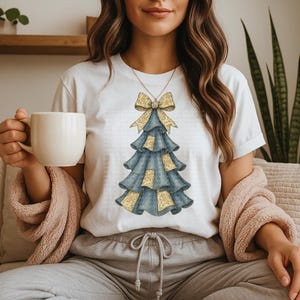 May include: White t-shirt featuring a Christmas tree design. The tree is blue with yellow floral accents and a yellow bow at the top. The person is holding a white mug and wearing a pink sweater.