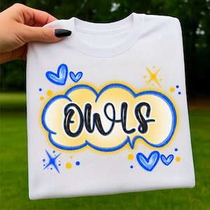 Puede incluir: Sudadera blanca con la palabra "OWLS" en escritura negra dentro de un diseño de nube amarilla y azul. El diseño incluye corazones azules y estrellas amarillas. La sudadera se muestra sobre un fondo verde.