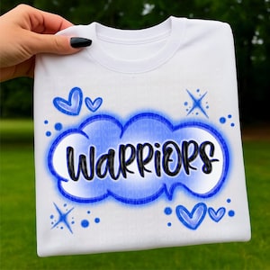Puede incluir: Camiseta blanca con la palabra "WARRIORS" en negro, delineada en azul, dentro de un diseño de nube azul. La camiseta está decorada con corazones azules y estrellas. La camiseta está sujeta por una mano.