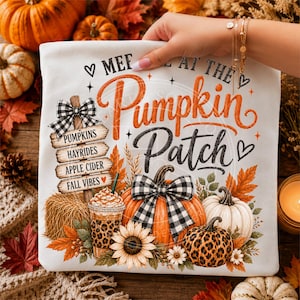 May include: A white t-shirt featuring a graphic print with the text Meet at the Pumpkin Patch. The design includes a wooden signpost listing Pumpkins, Hayrides, Apple Cider, and Fall Vibes, alongside pumpkins, a sunflower, and a leopard print cup.