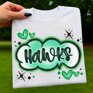 Puede incluir: Camiseta blanca con la palabra "Hawks" en letras negras dentro de un diseño de nube verde y negra. Corazones verdes y estrellas adornan el diseño de la camiseta.