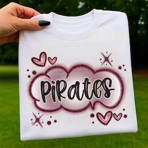 Puede incluir: Sudadera blanca con la palabra "Pirates" en un diseño de burbuja, adornada con corazones y estrellas. El texto es negro con un contorno granate y un relleno blanco rosado. El diseño incluye corazones rosas y estrellas.