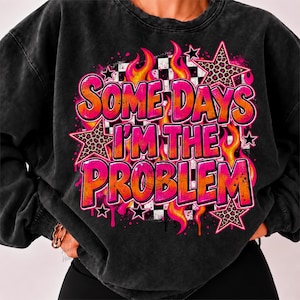 Puede incluir: Una sudadera negra con lavado ácido que presenta un estampado gráfico con el texto "SOME DAYS I'M THE PROBLEM" en letras con degradado rosa y naranja. El diseño incluye llamas naranjas, un fondo de cuadros blanco y negro y estrellas de leopardo.