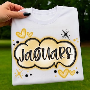 Puede incluir: Sudadera blanca con la palabra "Jaguars" en un diseño de burbuja negro y dorado. El diseño incluye corazones dorados, estrellas y puntos negros. La sudadera es sostenida por una mano con una uña pintada de negro.