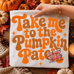 May include: A white t-shirt featuring the orange text Take me to the Pumpkin Patch in a retro font, accented by small orange star shapes. A small logo in the bottom right corner reads Heart & Soul Co. Original Designer in black and pink.