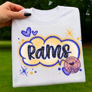 Puede incluir: Camiseta blanca con la palabra "Rams" en un diseño de nube azul y amarilla. La camiseta tiene cuello redondo y mangas cortas. Gráficos adicionales incluyen corazones, estrellas y un logotipo circular que dice "Original Heart & Soul Co Designer."