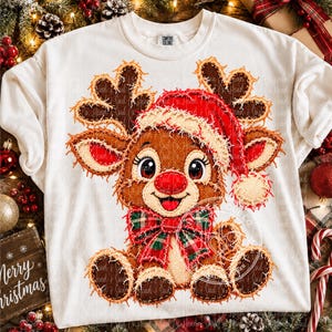 Può includere: Una t-shirt bianca a girocollo con la grafica di una renna marrone dal naso rosso, che indossa un cappello da Babbo Natale rosso e un papillon a quadri rossi e verdi. Il design ha un aspetto sfilacciato e testurizzato. Etichetta Comfort Colors.