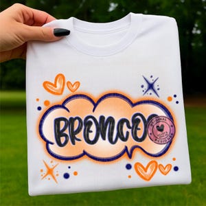 Könnte beinhalten: Weißes Sweatshirt mit dem Wort "BRONCO" in einem wolkenförmigen Design. Das Design umfasst orangefarbene und blaue Akzente, Herzen und Sterne. Ein kleines rundes Logo mit Text ist ebenfalls sichtbar.