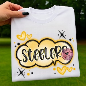 Puede incluir: Sudadera blanca con la palabra "Steelers" en letras negras, dentro de un diseño de nube amarilla y negra. El diseño incluye corazones amarillos, estrellas y un logotipo en forma de corazón rosa. La sudadera es sostenida por una persona.