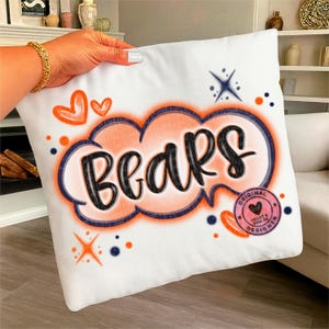 Puede incluir: Cojín cuadrado blanco con la palabra "BEARS" en escritura negra dentro de un diseño en forma de nube naranja y azul. El cojín también presenta corazones y estrellas naranjas. Un logotipo circular rosa con texto se encuentra en la esquina inferior derecha.
