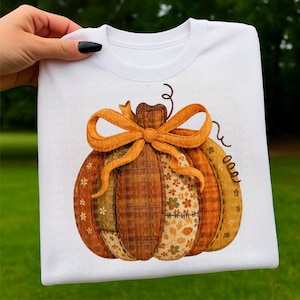 Op de afbeelding: Witte sweatshirt met een kleurrijk pompoenontwerp. De pompoen is gemaakt van verschillende stoffen lappen in bruin, oranje en geel, met een grote oranje strik. Het ontwerp heeft een herfstthema.