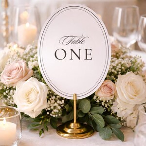 May include: An oval table number card with the words "Table ONE" in elegant script. The card is set on a gold stand, surrounded by a floral arrangement of white and blush roses, greenery, and baby's breath. Soft lighting and candles create a romantic ambiance.