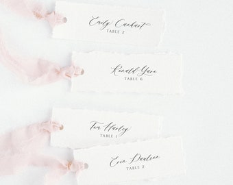 May include: Four white place cards with handwritten names and table numbers. The cards have a rustic look, with torn edges. The cards are tied with pink ribbon.