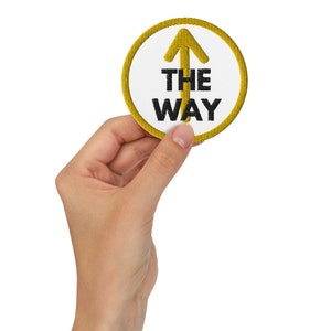 Op de afbeelding: Een gele cirkel met een witte achtergrond en een zwarte pijl die omhoog wijst. De tekst "THE WAY" staat in zwarte letters onder de pijl.