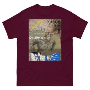 May include: A maroon t-shirt with a graphic design featuring a man with tattoos, a scallop shell, and the text "One Step at a Time". The graphic also includes the text "Santiago de Compostela".