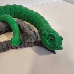 May include: Three toy snakes, one green and two gray, are curled together. The green snake has a textured surface and a detailed head with eyes. The gray snakes have smooth surfaces and detailed heads with eyes.