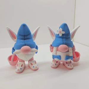 May include: Two Easter gnomes with bunny ears, one wearing a blue hat with a white flower and a blue bow, the other wearing a blue hat and holding a pink basket.