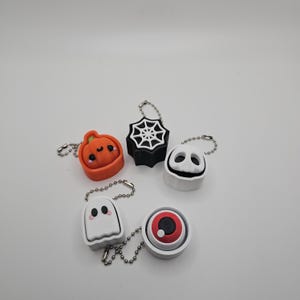 May include: A collection of Halloween-themed keychains. The keychains include a smiling orange pumpkin, a black spiderweb, a white ghost, a white face with black eyes and mouth, and a red and white eyeball. Each keychain has a silver chain.