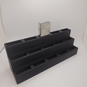 May include: A black, tiered display stand with multiple rectangular compartments. A silver-colored lighter sits on the top tier. The stand is designed to showcase small items, likely for retail or personal organization.