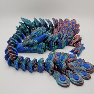 May include: A 3D printed dragon figurine with a peacock-inspired design. The dragon is curled up in a sleeping position with its head resting on its tail. The scales are a mix of blue, green, and copper colors.
