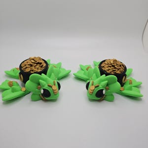 May include: Two green frog figurines with black pots filled with gold coins on their backs. The frogs have black eyes and gold accents.