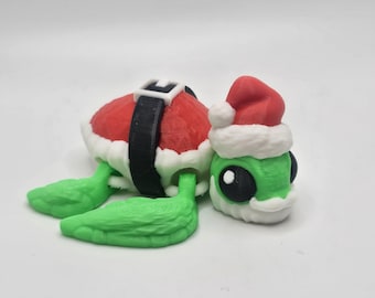 May include: A 3D-printed Christmas-themed turtle figurine. The turtle has a red shell with a white trim and a black belt. It has green flipper-like legs and a green head wearing a Santa hat. The figurine is on a white background.