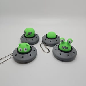 May include: Four novelty keychains featuring alien-themed designs. Each keychain has a gray, saucer-shaped base with a green alien figure on top. One alien has large eyes, another has a classic alien face, and the others have unique designs. Each keychain has a silver chain.