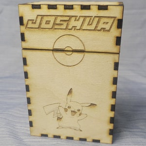 May include: A wooden box with a black border and the name "Joshua" engraved on the top. The box features a Pokemon character, Pikachu, and a Pokeball symbol.