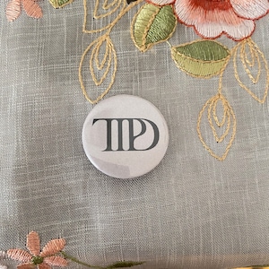 May include: A round, light gray button with the letters "TIP" in dark gray. The button is set against a gray, textured background with floral embroidery in shades of pink, green, and beige.