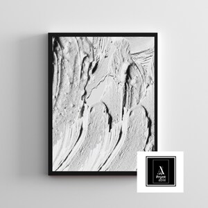 May include: A framed abstract art print featuring a textured, monochromatic white paint design. The artwork has a black frame and a logo in the bottom right corner. The paint strokes create a dynamic, layered effect.