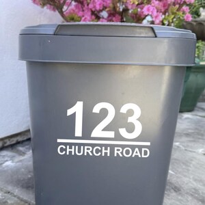 May include: A grey outdoor bin with the address "123 CHURCH ROAD" in white lettering. The bin is in front of a flowering bush with pink blossoms.