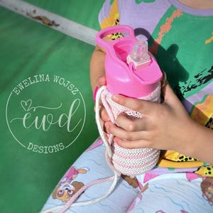 Könnte beinhalten: Eine rosa Wasserflasche mit einer weiß-rosa gestrickten Hülle und einem passenden Riemen. Die Flasche wird von einer Person gehalten, die eine hellblaue Shorts mit einem Cartoon-Hunde-Aufdruck und ein farbenfrohes Hemd trägt. Der Text "Ewelina Wojsz ewd DESIGNS" ist sichtbar.