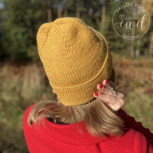 May include: A mustard yellow knitted beanie with a folded brim. The beanie is worn by a person with long blonde hair and a red jacket. The text "Ewelina Wojsz ewd Designs" is visible in the upper right corner of the image.