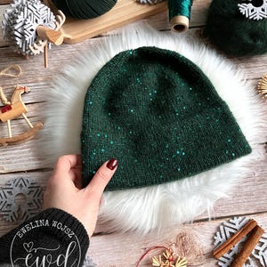 May include: A dark green knit beanie with scattered sequins, resting on white faux fur. The hat is surrounded by Christmas-themed decorations, including snowflakes, yarn, and cinnamon sticks. The beanie is held by a hand.