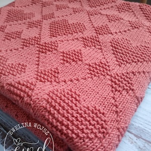 Puede incluir: Una manta tejida de color coral que presenta un patrón geométrico repetitivo de corazones y diamantes texturizados. La tela muestra un punto bobo consistente en todo el diseño. Un logotipo circular con el texto EWELINA WOJSZ ewd DESIGNS es visible.