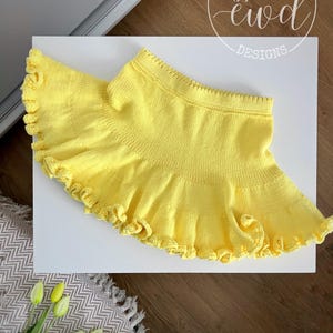 Pode incluir: Uma saia de malha amarela brilhante com bainha com babados. A saia é exibida em uma superfície branca. A imagem também inclui o texto "Ewelina Wojsz ewd DESIGNS" em um logotipo circular.