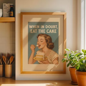 Puede incluir: Impresión enmarcada en madera con el texto "WHEN IN DOUBT, EAT THE CAKE" con una ilustración vintage de una mujer comiendo pastel. La impresión se muestra en un entorno de cocina con utensilios de madera y una tetera de acero inoxidable.