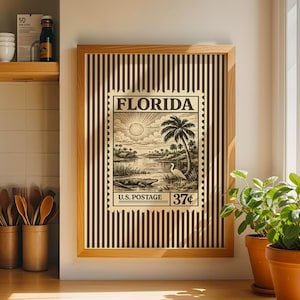 Op de afbeelding: Een ingelijste print met een vintage Florida postzegelontwerp. De postzegel toont een landschap met een zon, palmbomen en water. De lijst heeft een streepjespatroon. De print is gelabeld met "U.S. Postage 37¢".