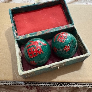 May include: A vintage green box with a red interior contains two green stone massage balls. Each ball features red Chinese characters and floral accents. The box is trimmed in gold and has a small clasp.