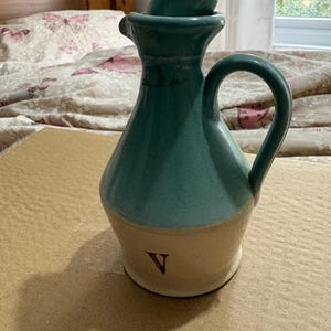 May include: A teal and cream ceramic jug with a handle and a small lid. The jug has a rounded body and a narrow neck. A letter "V" is painted on the base.