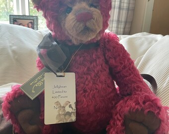 May include: A burgundy teddy bear with a light brown face and ears. The bear has a tag that reads "Jellybean Limited to 400 Pieces No. 182." It is sitting on a brown bag with gold lettering. A black ribbon is tied around its neck.