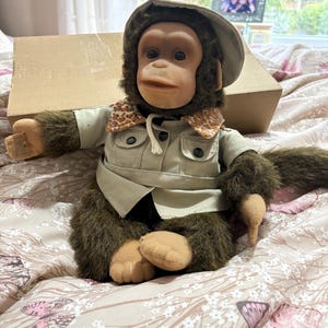 May include: A plush monkey doll wearing a khaki safari-style jacket and hat. The monkey has brown fur, a tan face, and is sitting on a floral patterned bedspread. A cardboard box is behind the doll.