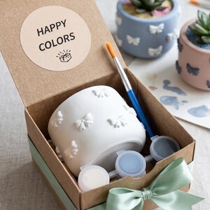 Può includere: Una scatola regalo in cartone marrone legata con un nastro verde salvia contenente un vaso in ceramica bianca con dettagli a farfalla in rilievo, un pennello blu e tre piccoli vasetti di vernice. Il coperchio ha un adesivo HAPPY COLORS.